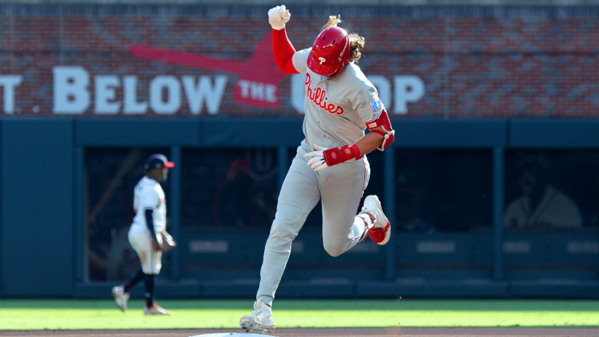 Phillies player in a gray uniform raises a fist while running, with another player blurred in the background.