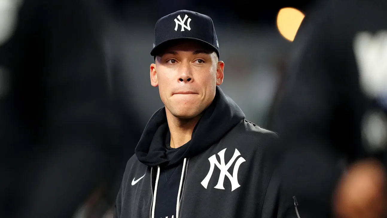 A man in a Yankees cap and black team jacket stands against a blurred stadium background.