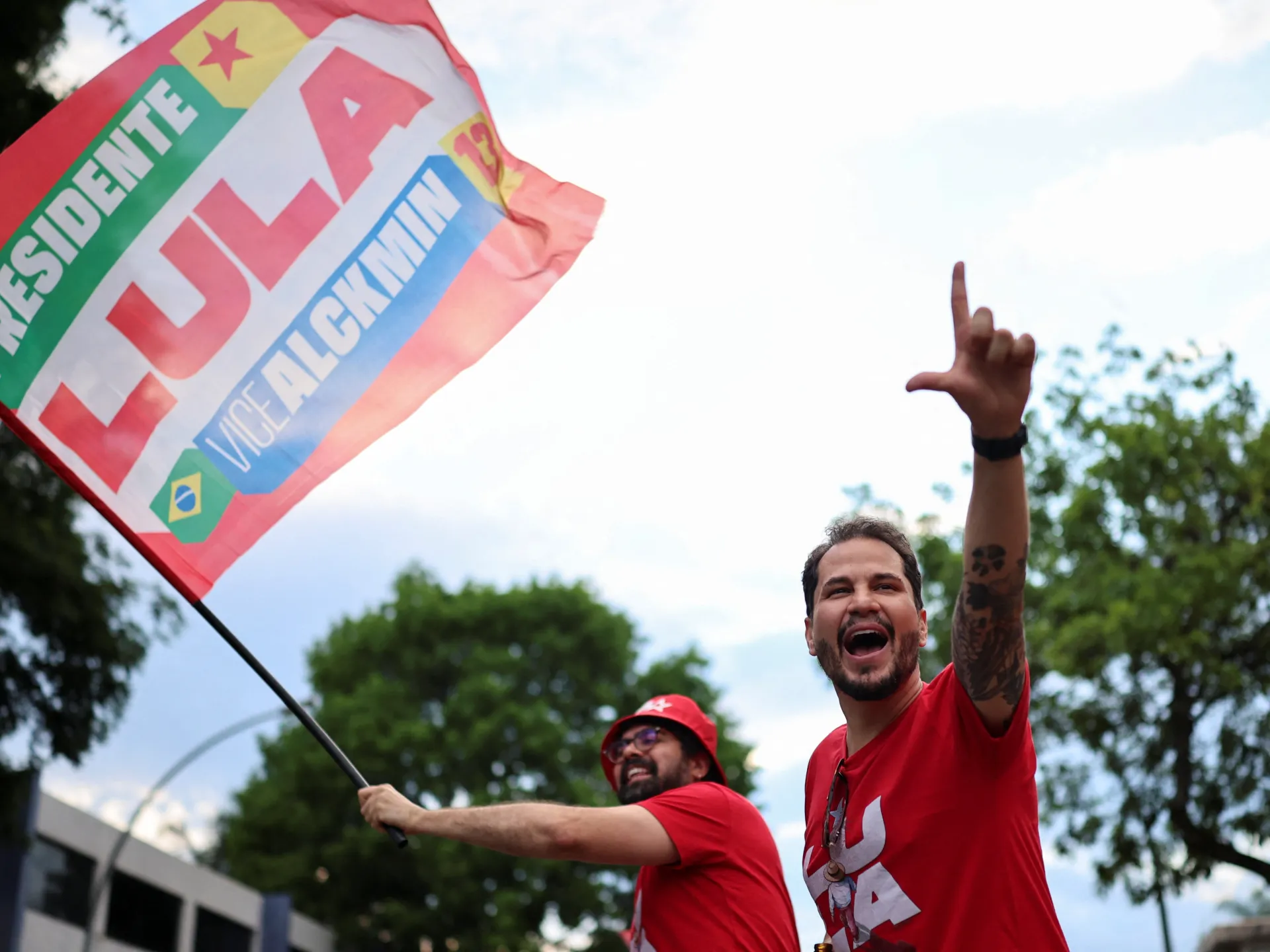 Two men in red shirts cheer as one waves a political flag and the other raises his hand.