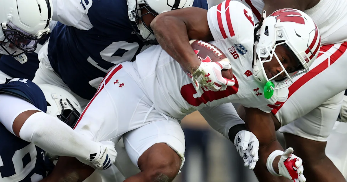 Wisconsin player in a white uniform carries the football as defenders in navy tackle him.