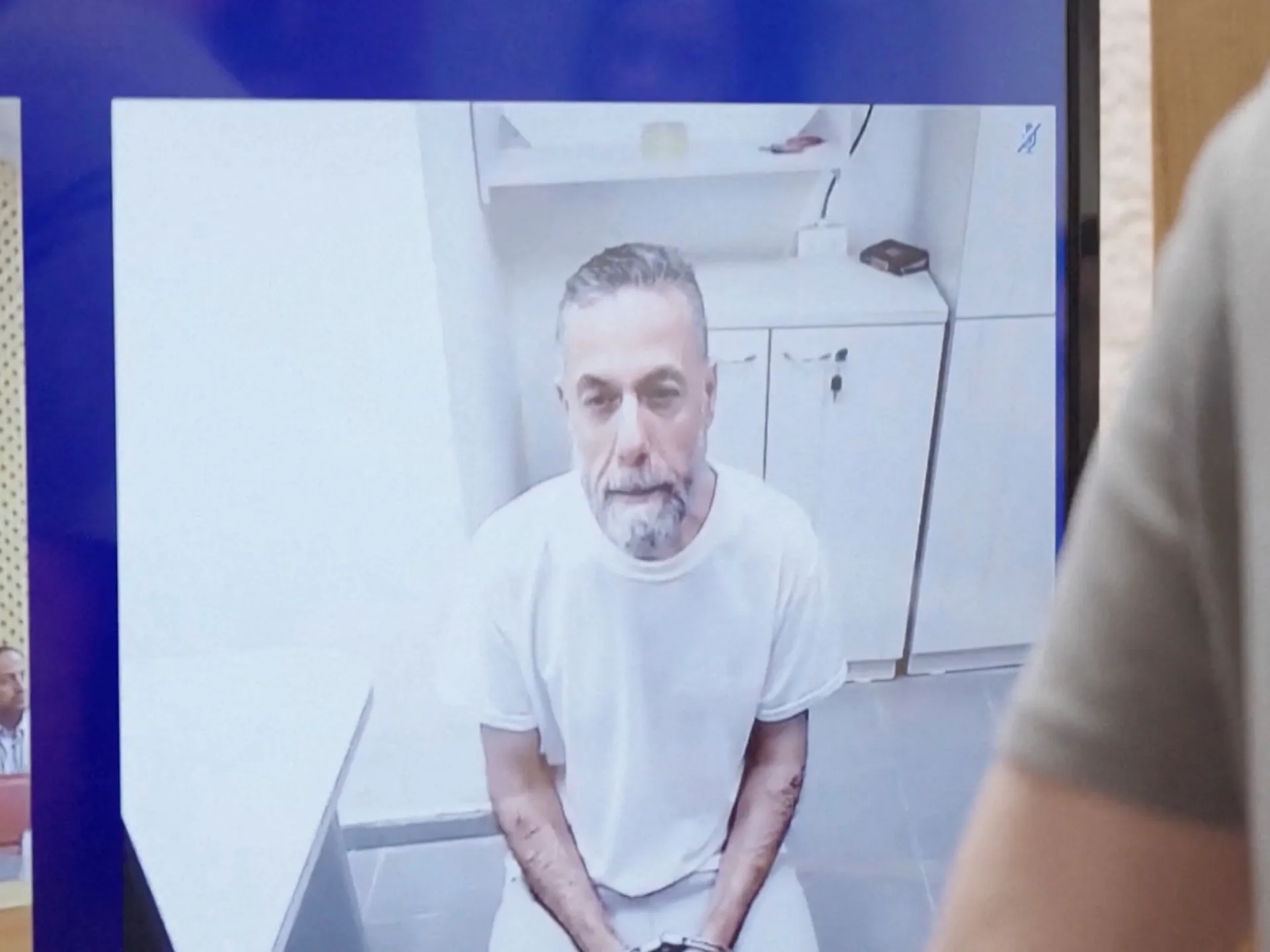 A gray-haired man in a white shirt appears on a video call, seated in a stark room.