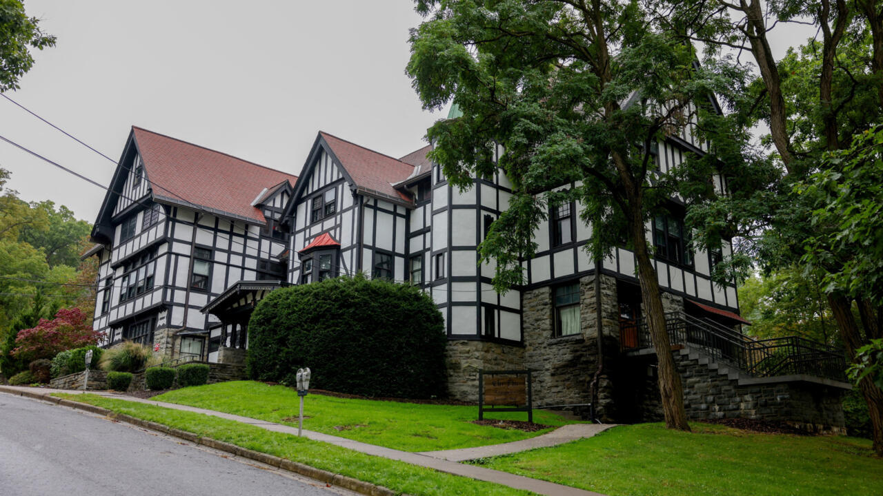 Tudor-style building with dark timber framing, stone walls, red roofs, and mature trees beside a street.