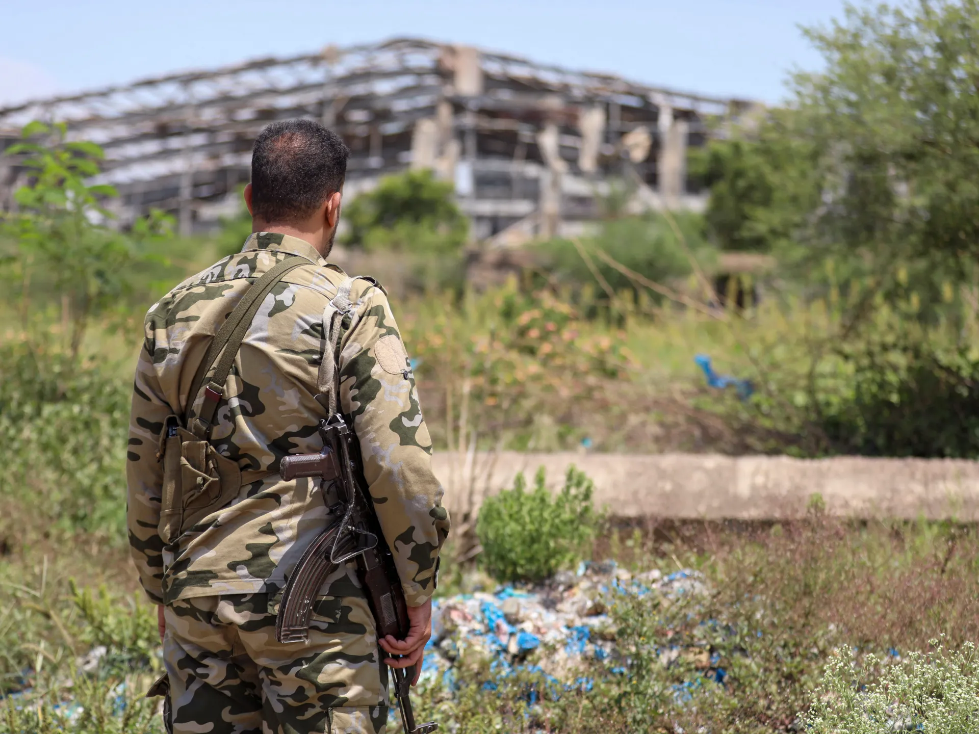 A uniformed man with a rifle stands amid brush, facing a damaged structure in the distance.
