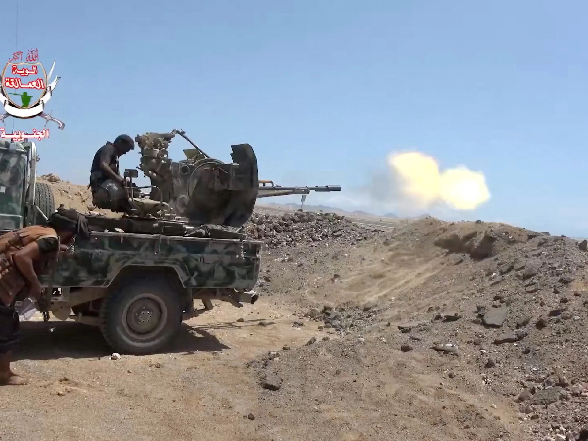 A mounted gun fires from a camouflage truck on rocky terrain, with two men beside it.