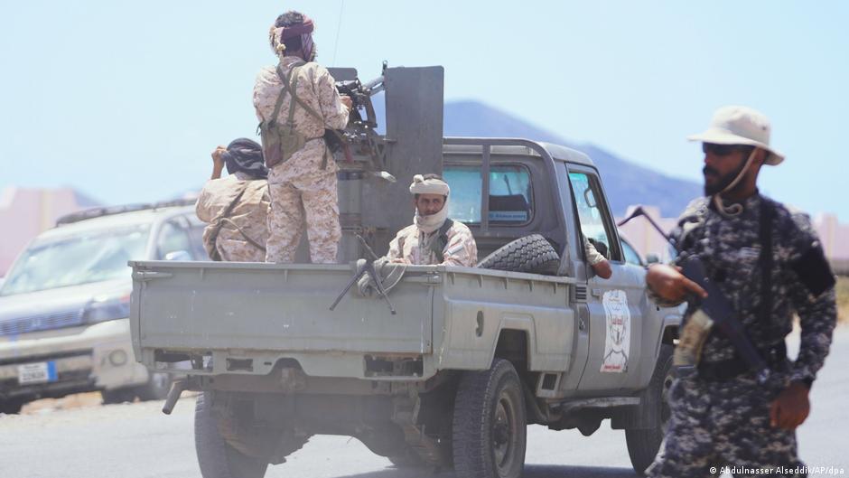 Armed men ride in the back of a pickup truck on a sunlit road.