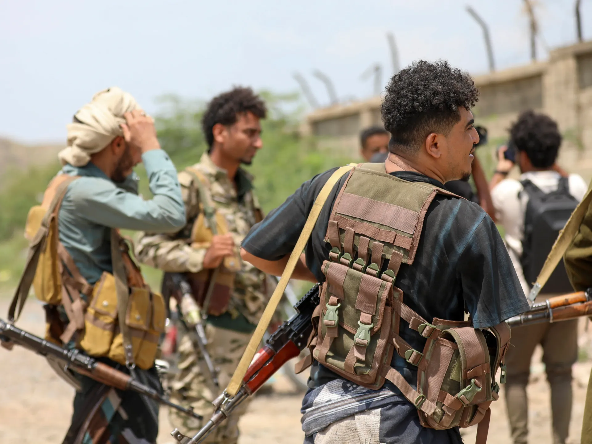 Armed men in tactical gear stand outdoors, with rifles and a concrete wall in the background.