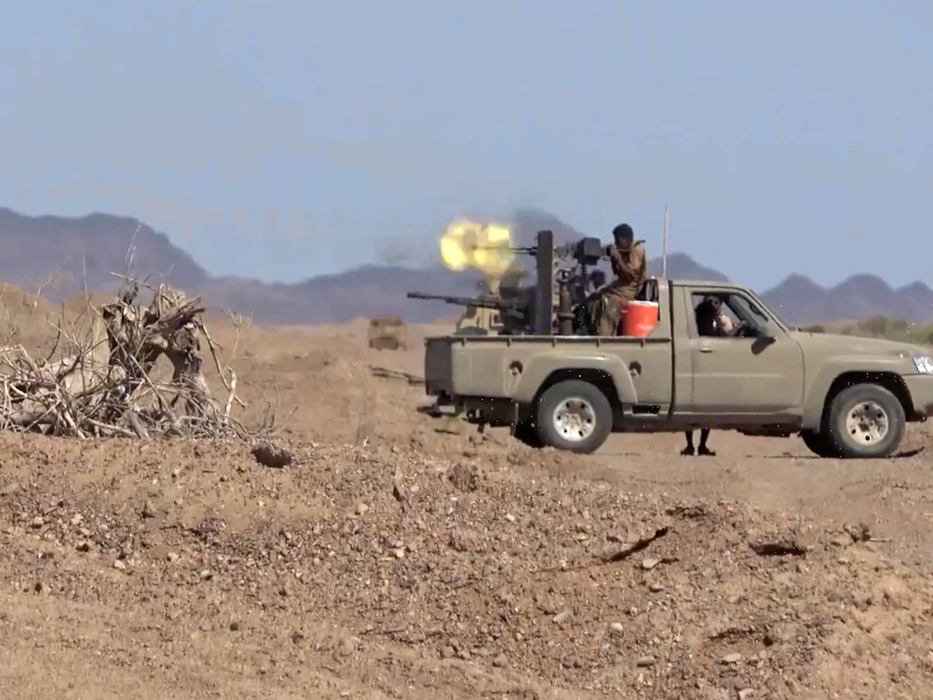 A pickup-mounted gun fires in a rocky desert as armed men ride in the truck bed.