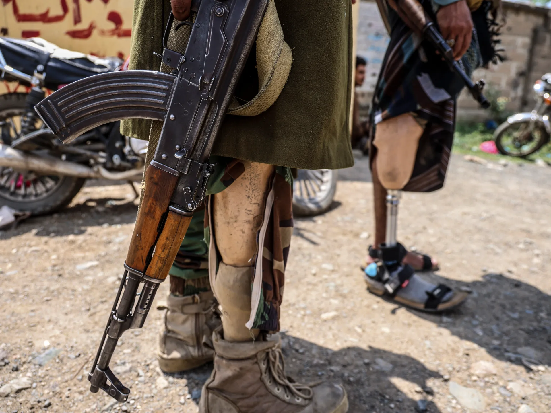 Armed men stand among motorcycles; one holds a rifle, while another wears a prosthetic lower leg.